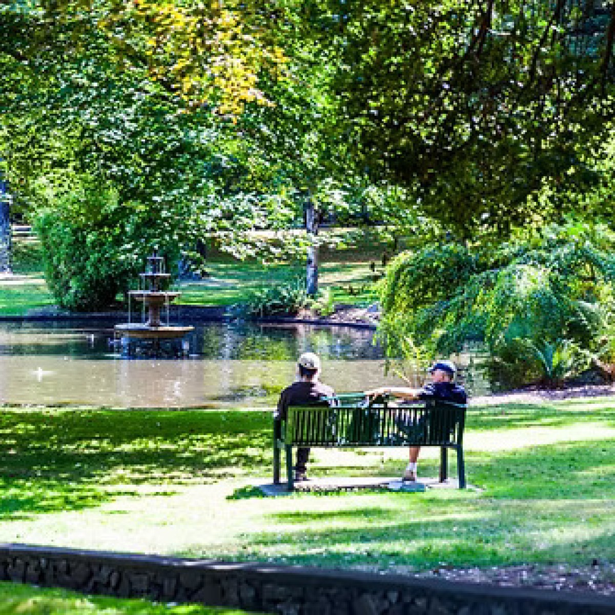 Two people sitting on a bench by a pond in a lush, green park.