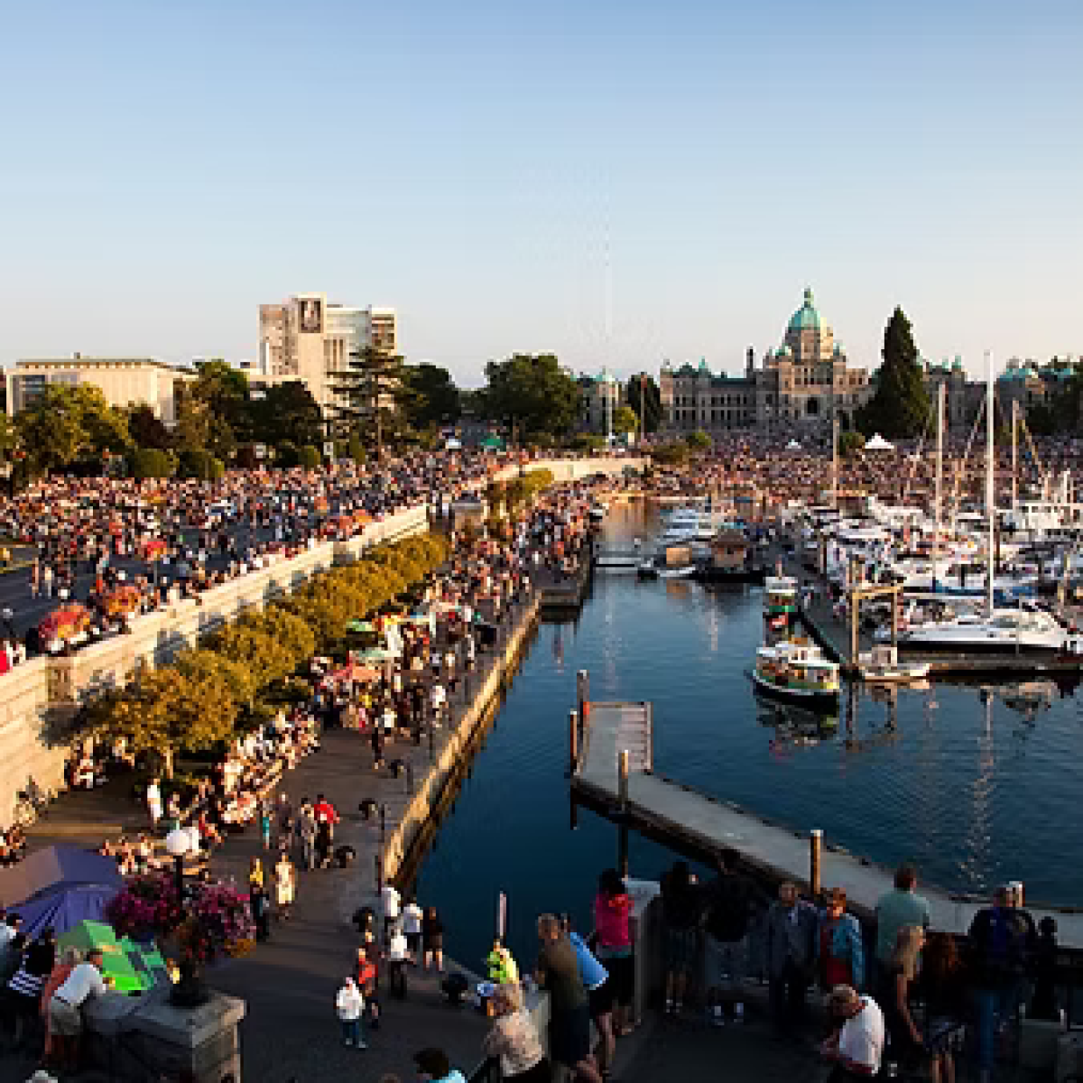 Crowd gathers near waterfront with boats and historic building in the background.